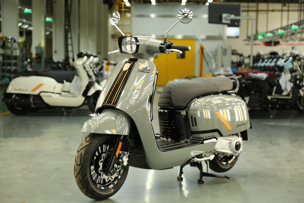 Ein silberner Royal Alloy Retro-Roller mit grauem Sitz und schwarzen Reifen steht in einem hellen Raum. Im Hintergrund sind weitere Roller zu sehen.